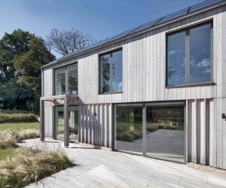 The rear elevation of a contemporary, timber-clad, self build home. Large sliding doors, from VELFAC, on the ground floor sit adjacent to a large patio and landscaped garden. The first floor features large windowsm and solar panels are installed on the roof.