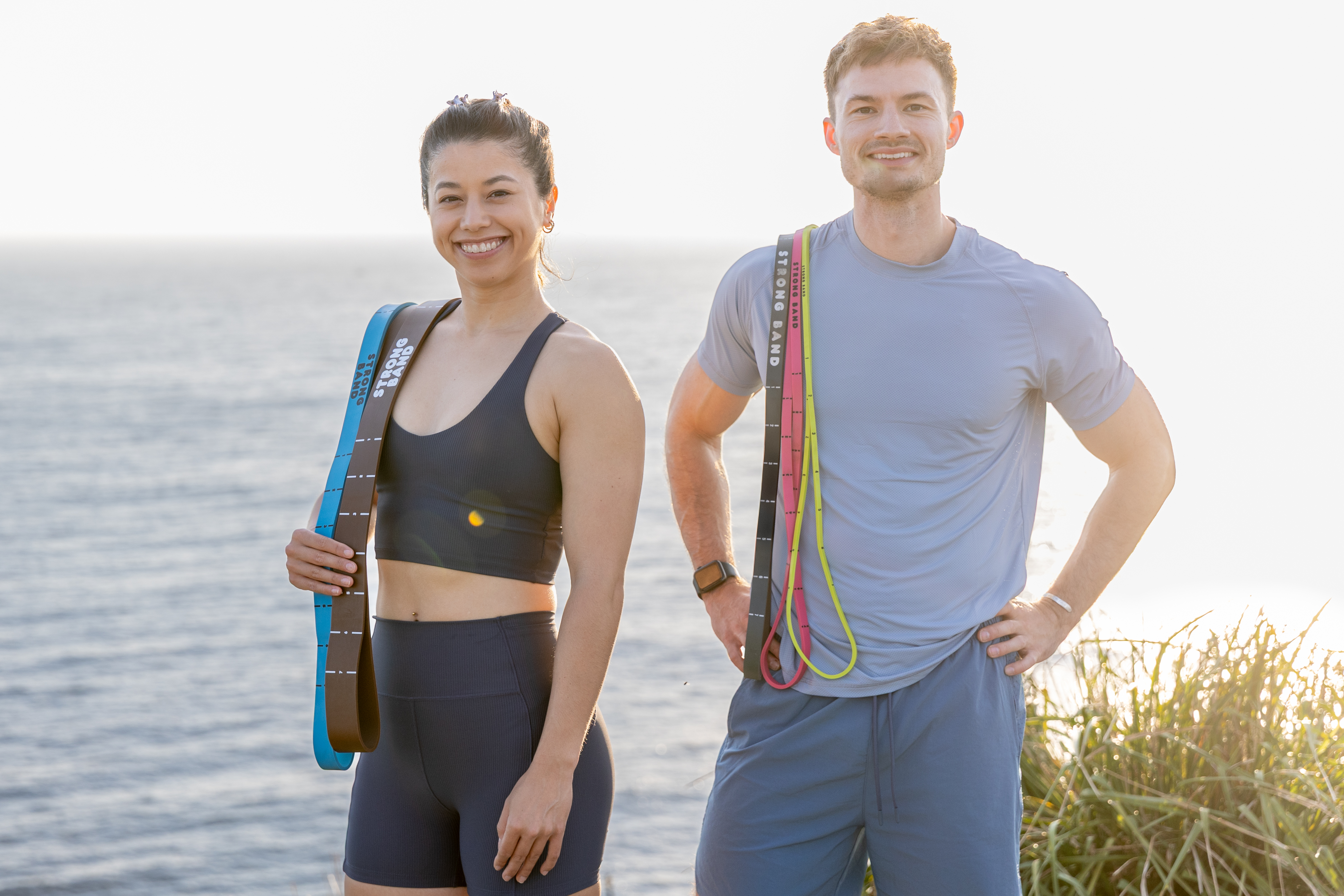 Man and woman stand outside with sea behind them. They have multiple resistance bands draped over one shoulder