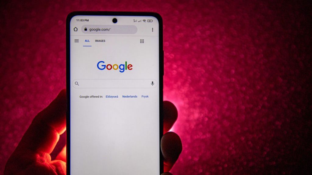 A phone showing the google homepage is held in someone&amp;#039;s hand in close-up, with a dimly-lit red wall in the background