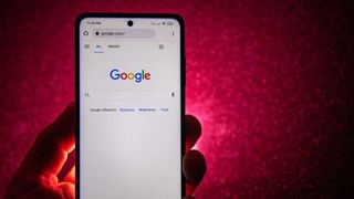 A phone showing the google homepage is held in someone's hand in close-up, with a dimly-lit red wall in the background
