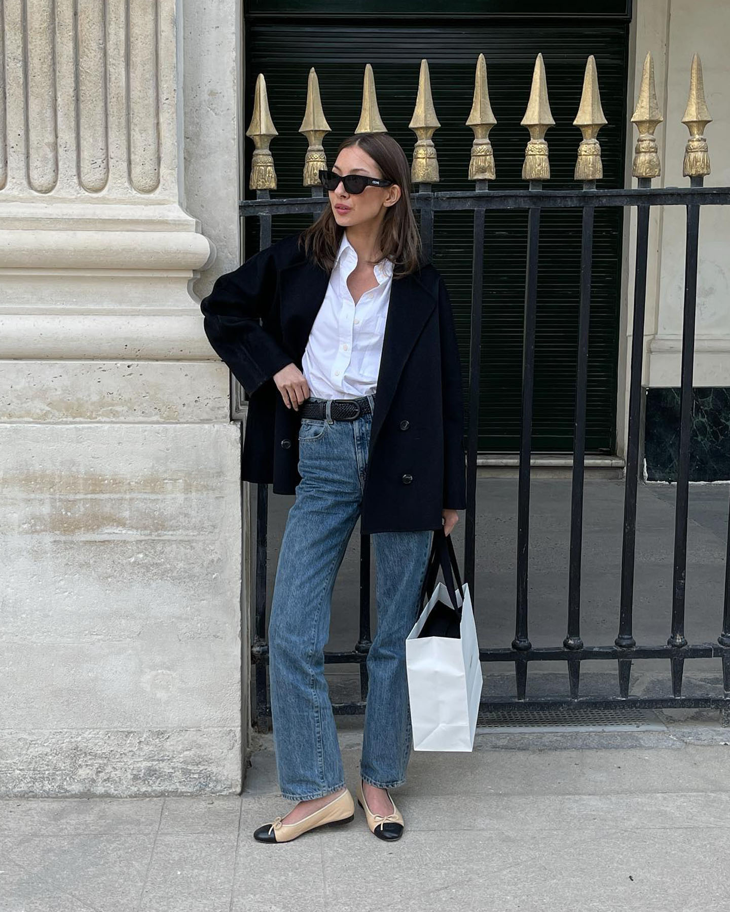 fashion influencer Felicia Akerstrom poses on the sidewalk in Paris wearing a black blazer, white button-down shirt, belt, jeans, and Chanel cap-toe ballet flats