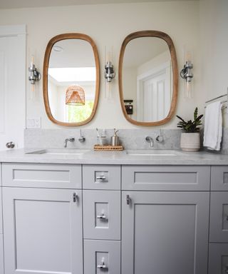 Two wooden rectangular curved mirrors with three silver chrome wall sconces and a gray sink unit with two faucets and two toothbrush holders on a rattan tray, with a towel rail and a white towel to the right