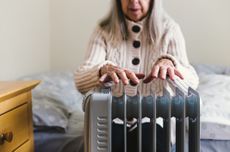 A cold-looking older woman puts her hands over the top of a radiator