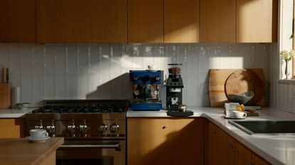 Image of a navy blue La Marzocco machine on a white kitchen countertop with wooden cabinetry. There is a black coffee grinder and scale next to it.