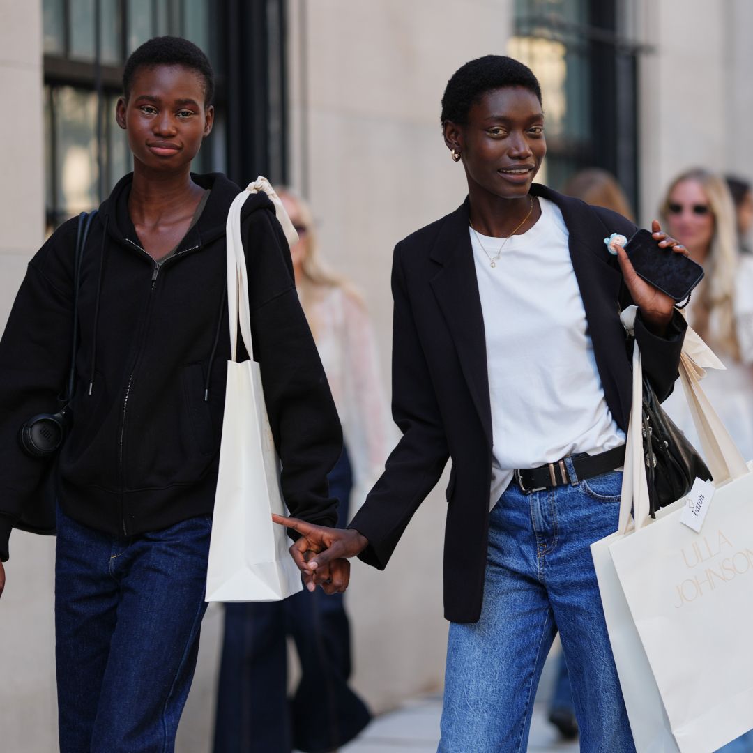 A guest (L) wears black zipper high neck sweater, a black shiny leather shoulder bag, navy blue denim flared pants, a white oversized shoulder shopping bag, a guest (R) wears short natural hair, gold hoop earrings and a thin gold necklace, carries a black leather shoulder bag and a white paper shopping bag printed with ULLA JOHNSON, a black tailored blazer jacket over a white crew-neck T-shirt tucked into medium-blue high-waisted straight-leg denim jeans secured with a black leather belt with a silver buckle, outside Ulla Johnson, during New York Fashion Week, on September 14, 2025 in New York, New York