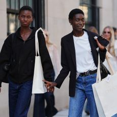 A guest (L) wears black zipper high neck sweater, a black shiny leather shoulder bag, navy blue denim flared pants, a white oversized shoulder shopping bag, a guest (R) wears short natural hair, gold hoop earrings and a thin gold necklace, carries a black leather shoulder bag and a white paper shopping bag printed with ULLA JOHNSON, a black tailored blazer jacket over a white crew-neck T-shirt tucked into medium-blue high-waisted straight-leg denim jeans secured with a black leather belt with a silver buckle, outside Ulla Johnson, during New York Fashion Week, on September 14, 2025 in New York, New York