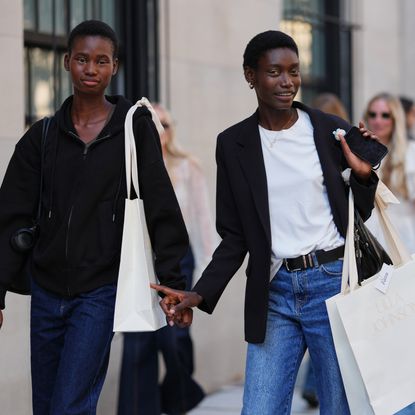 A guest (L) wears black zipper high neck sweater, a black shiny leather shoulder bag, navy blue denim flared pants, a white oversized shoulder shopping bag, a guest (R) wears short natural hair, gold hoop earrings and a thin gold necklace, carries a black leather shoulder bag and a white paper shopping bag printed with ULLA JOHNSON, a black tailored blazer jacket over a white crew-neck T-shirt tucked into medium-blue high-waisted straight-leg denim jeans secured with a black leather belt with a silver buckle, outside Ulla Johnson, during New York Fashion Week, on September 14, 2025 in New York, New York