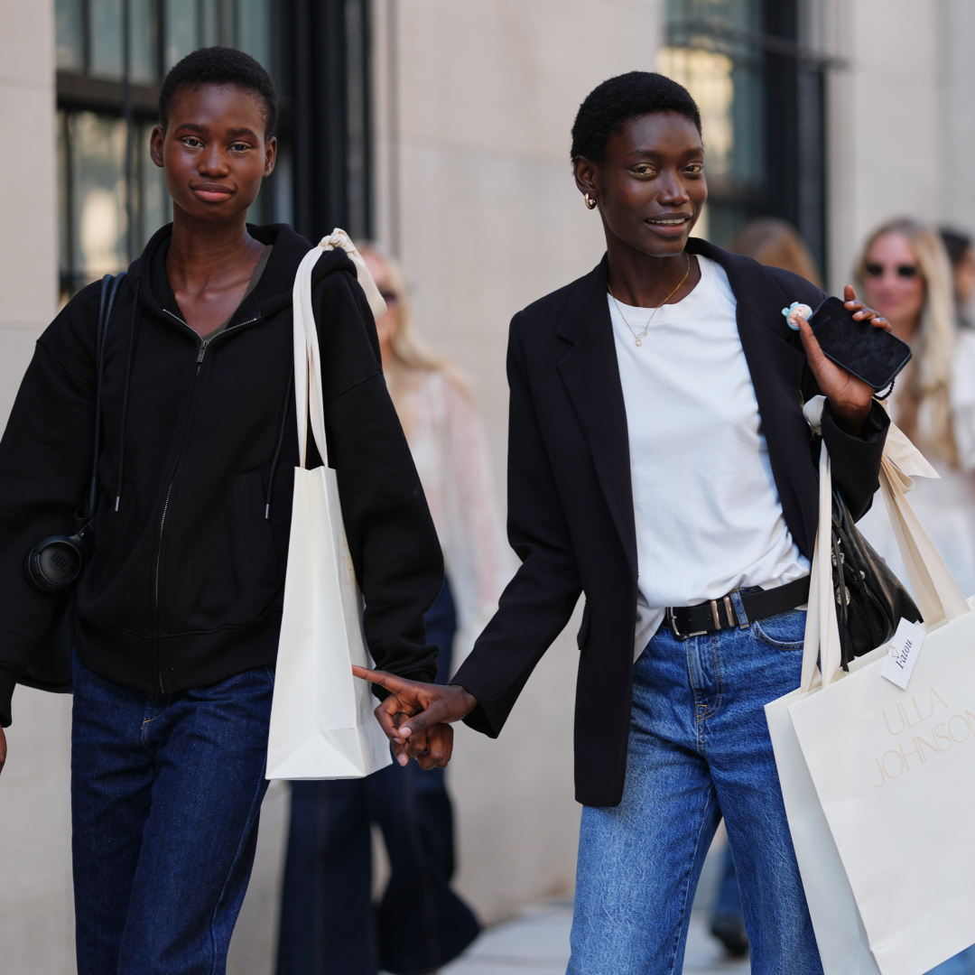 A guest (L) wears black zipper high neck sweater, a black shiny leather shoulder bag, navy blue denim flared pants, a white oversized shoulder shopping bag, a guest (R) wears short natural hair, gold hoop earrings and a thin gold necklace, carries a black leather shoulder bag and a white paper shopping bag printed with ULLA JOHNSON, a black tailored blazer jacket over a white crew-neck T-shirt tucked into medium-blue high-waisted straight-leg denim jeans secured with a black leather belt with a silver buckle, outside Ulla Johnson, during New York Fashion Week, on September 14, 2025 in New York, New York