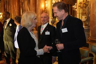 Queen camilla wearing a black-and-white dress and shaking hands with actor Damian Lewis, who is wearing a black suit and holding a wine glass