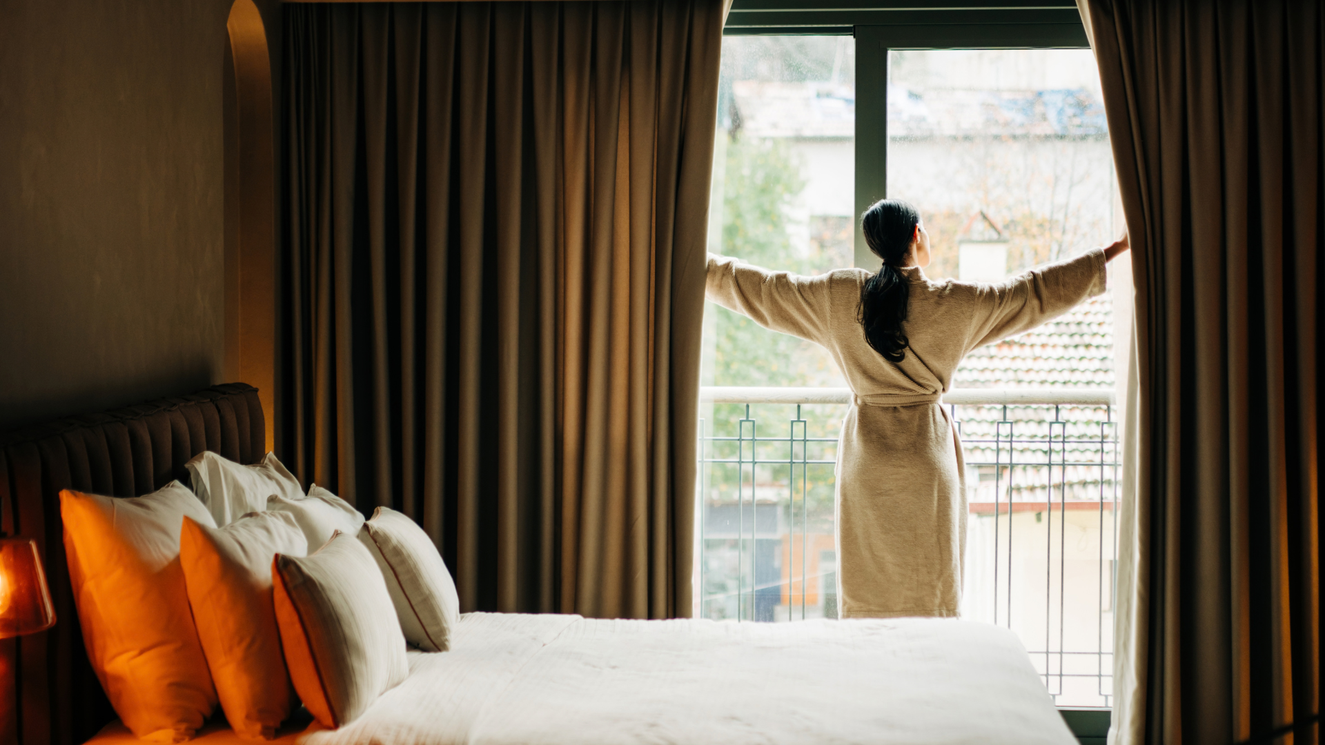 A woman opens the curtains of her bedroom in the morning to let the sunlight in.