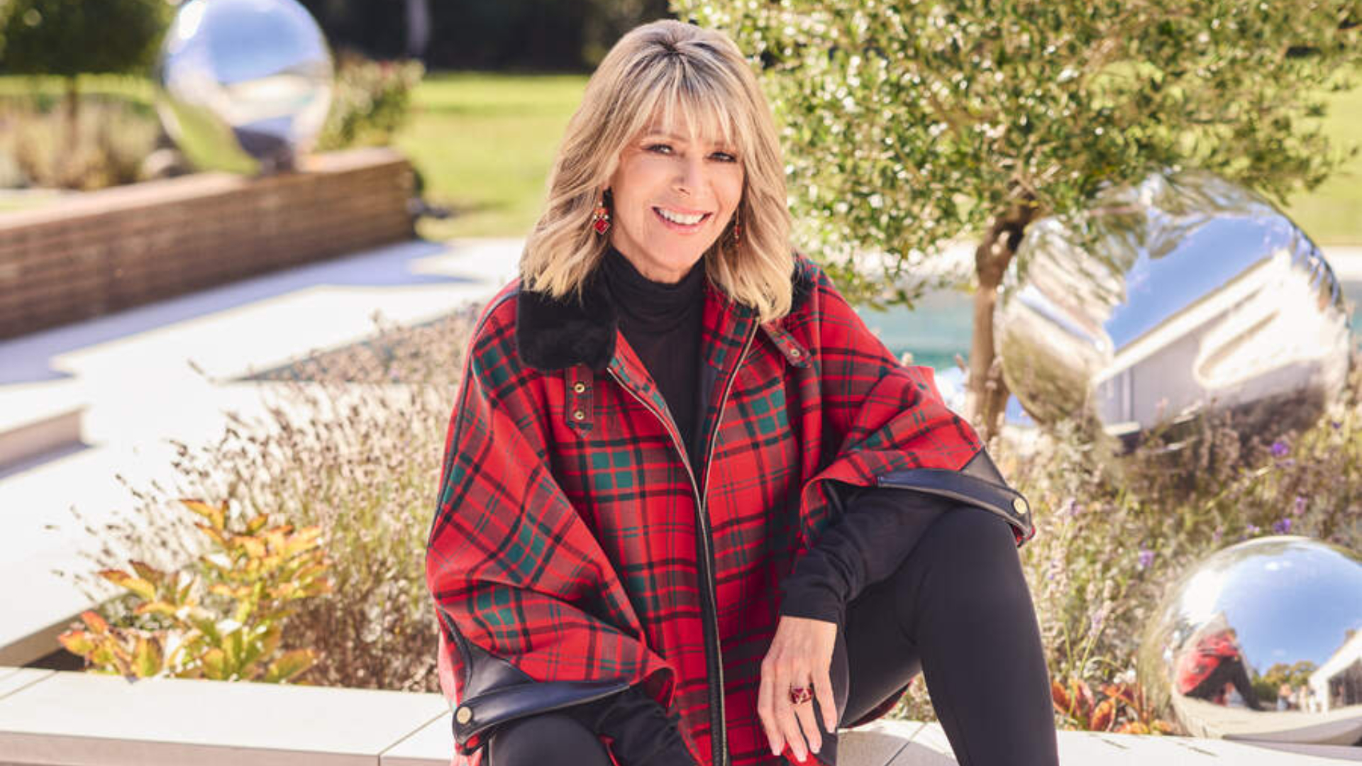 Ruth Langsford wearing a red checked jacket and boots in a garden and smiling at the camera
