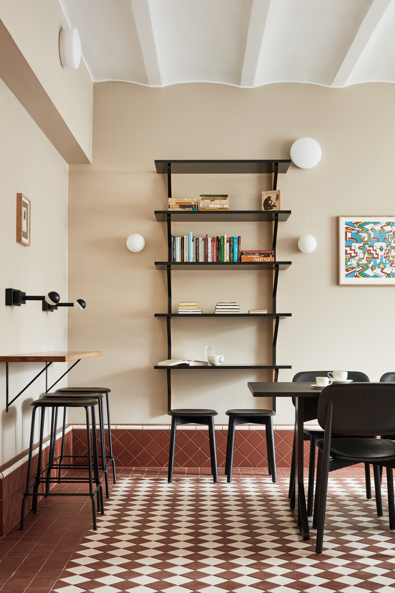 A checkered-floor hotel cafe features red and white tiling, buttery walls, wooden shelves painted back with matching tables, quirky artwork, and spherical lighting.