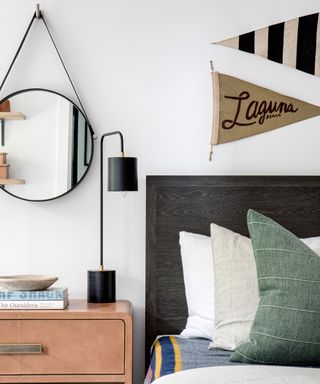 Close up of side of bed, small flag wall decorations, rounded mirror above wooden bedside table, table decorated with black table lamp, books and decorative bowl