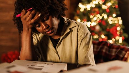 Young woman sitting her desk with a Christmas tree in the background, head in hand, contemplating her finances during the holidays.