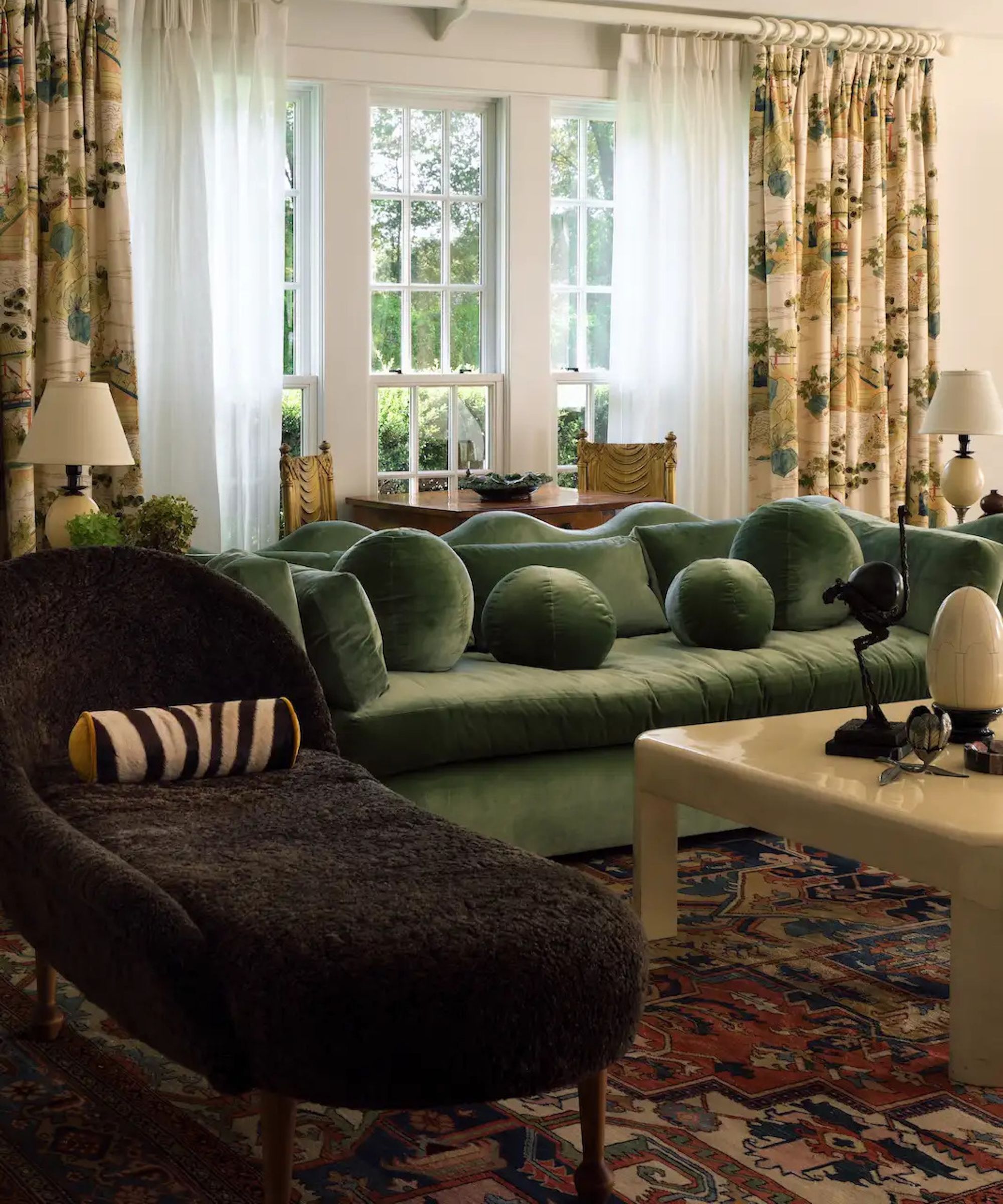 neutral living room with large windows, floral curtains, a green wavy velvet couch with ball pillows, a chaise lounge with lumbar pillow and lacquered coffee table