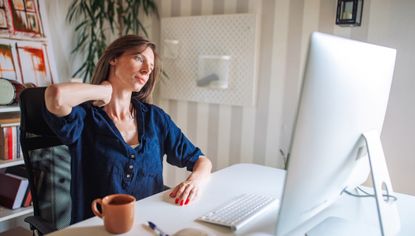 Woman sitting at a desk looking at a computer screen. She leans her head to one side and presses her hand into the side of her neck