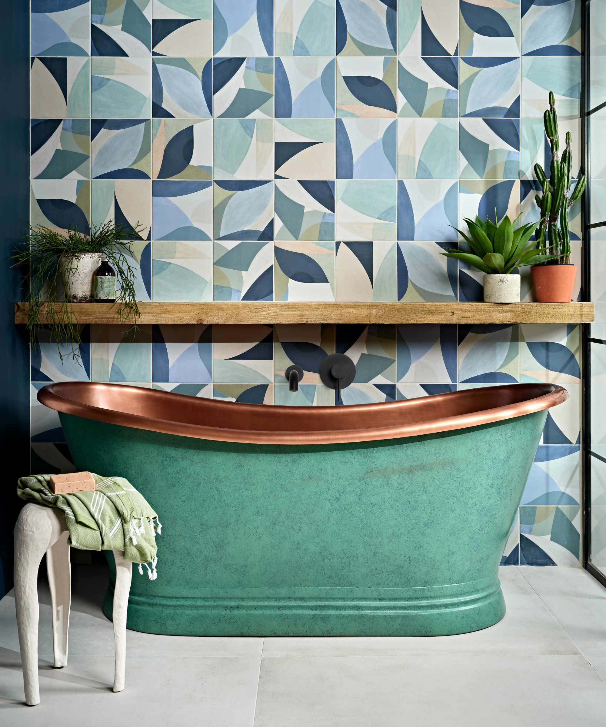 green and copper freestanding bath in bathroom with blue, green and cream geometric tiled wall and wooden shelf