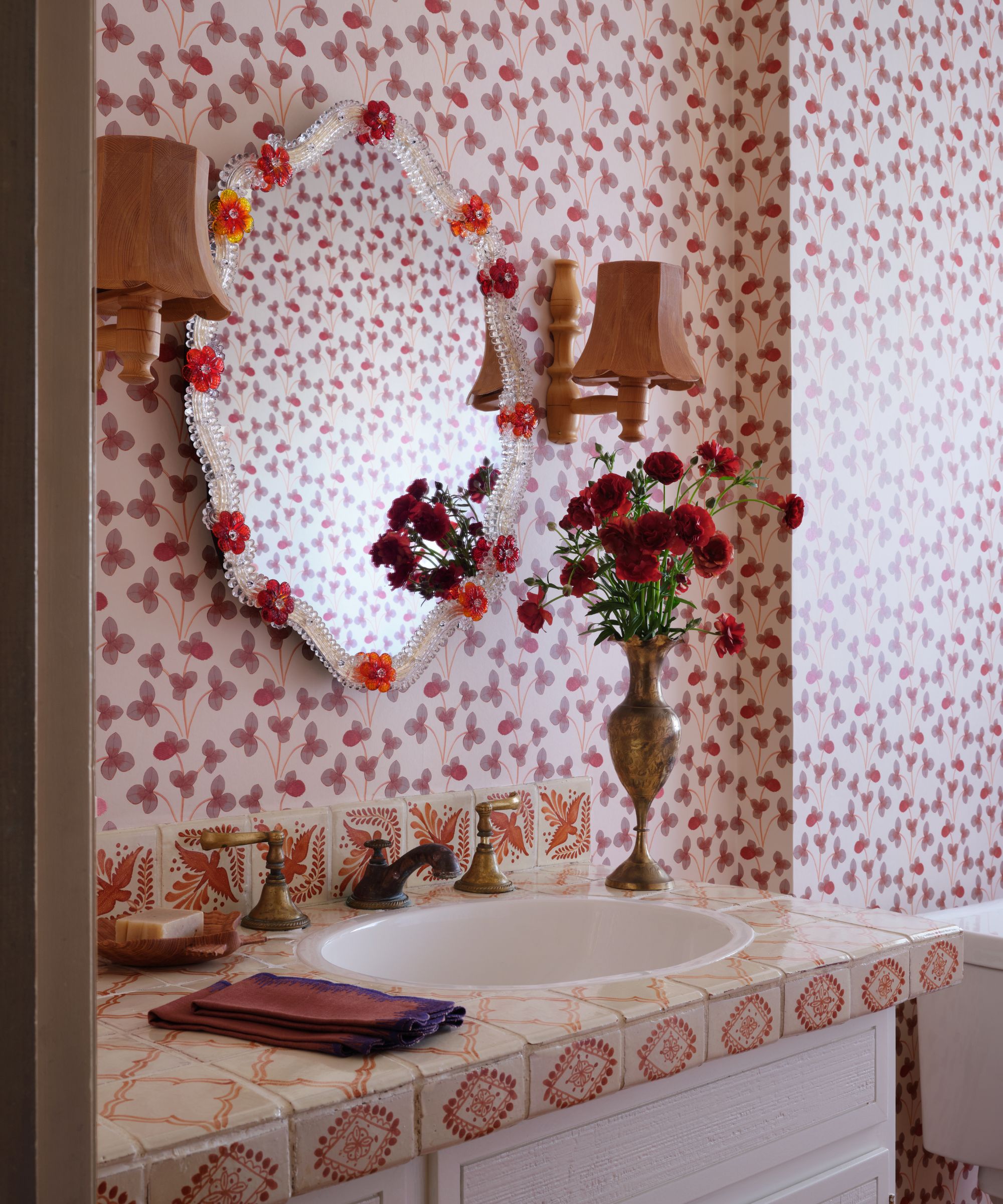 An angled view of a bathroom vanity featuring an ornate mirror decorated with glass flowers, a tiled countertop with painted patterns, and a brass vase of red flowers