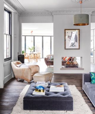 Living room with dark wooden floor, grey ottoman on a white rug next to a grey sofa and view through to the kitchen diner. Feature fireplace, with large brown leather lounge chair and footstool positioned beside