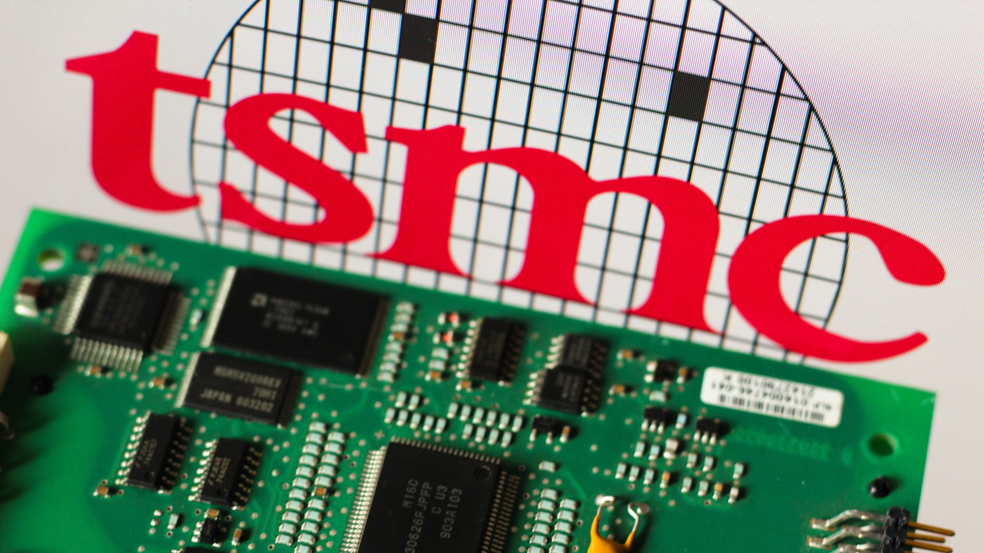 The TSMC (Taiwan Semiconductor Manufacturing Company) logo is seen in the background beside a printed circuit board (PCB).