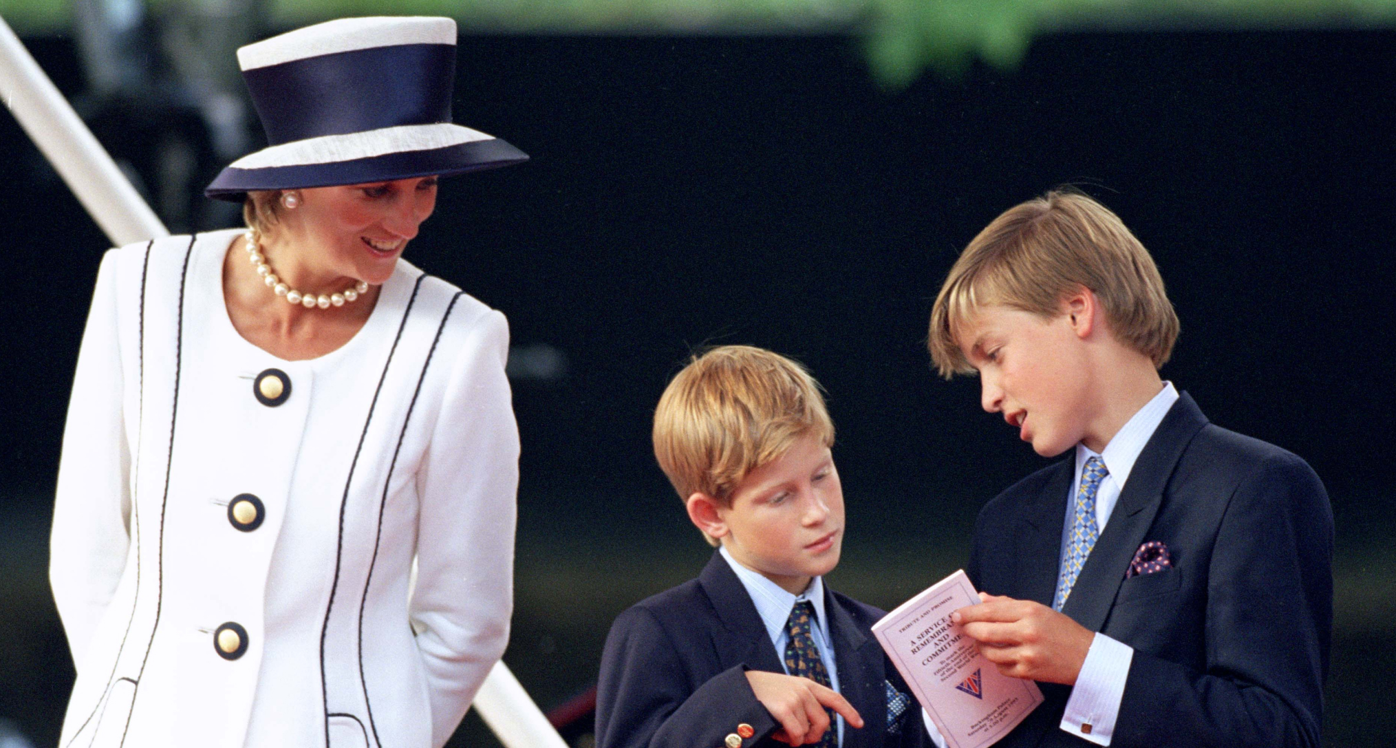 Princess Diana wearing a white suit and hat smiling at Prince Harry and Prince William, who are looking at a program