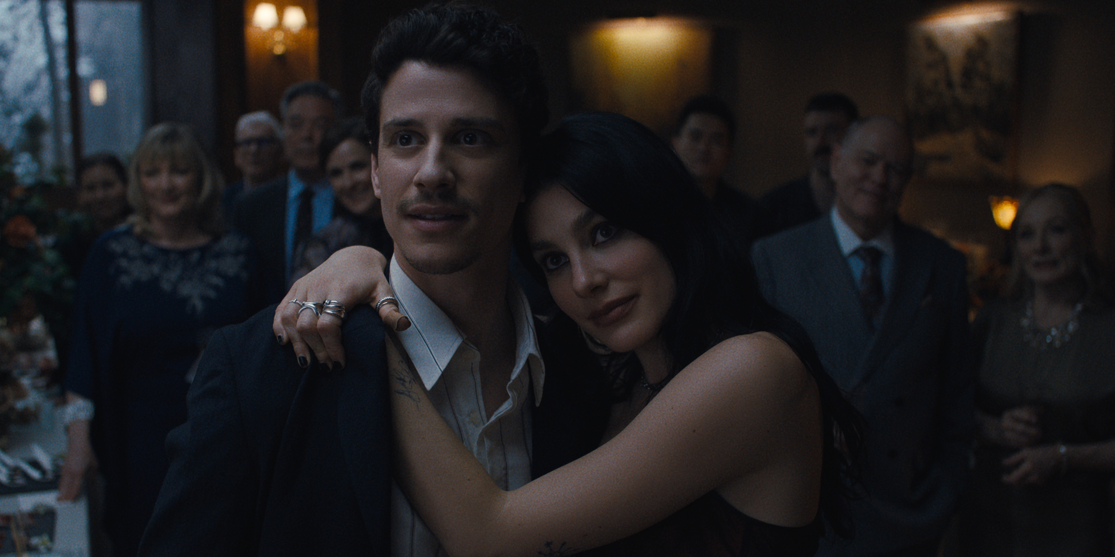 Adam DiMarco as Nicky Cunningham, Camila Morrone as Rachel Harkin in episode 105 of Something Very Bad Is Going To Happen wearing formal wear and holding each other while standing in front of a crowd at a party