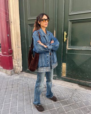 A woman stands in front of a doorway wearing denim on denim with brown clogs.