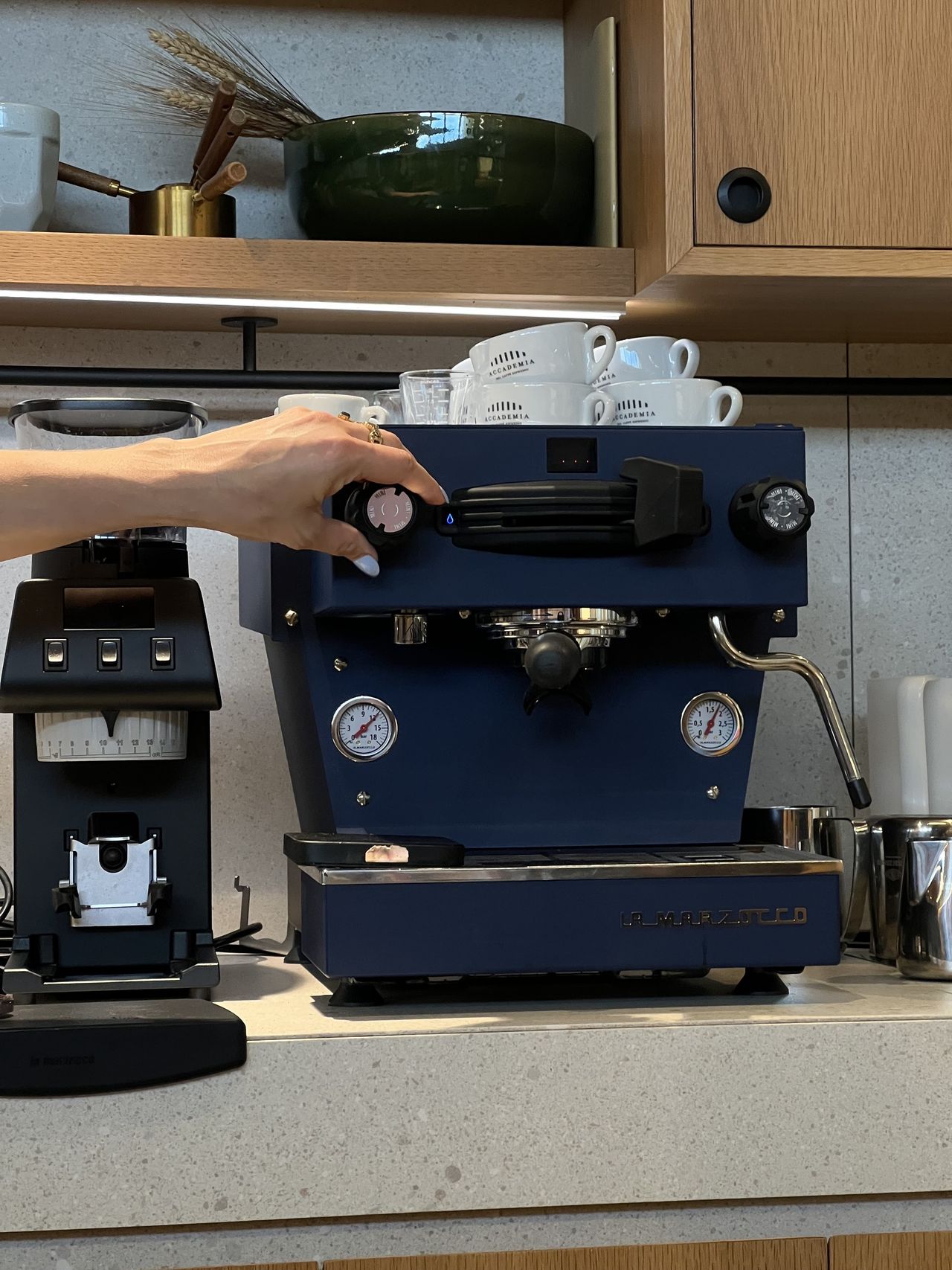 A hand turning a dial on a Navy Blue La Marzocco coffee machine with white coffee mugs on top of it and a black La Marzocco coffee grinder next to it. The machine is on a tan countertop.
