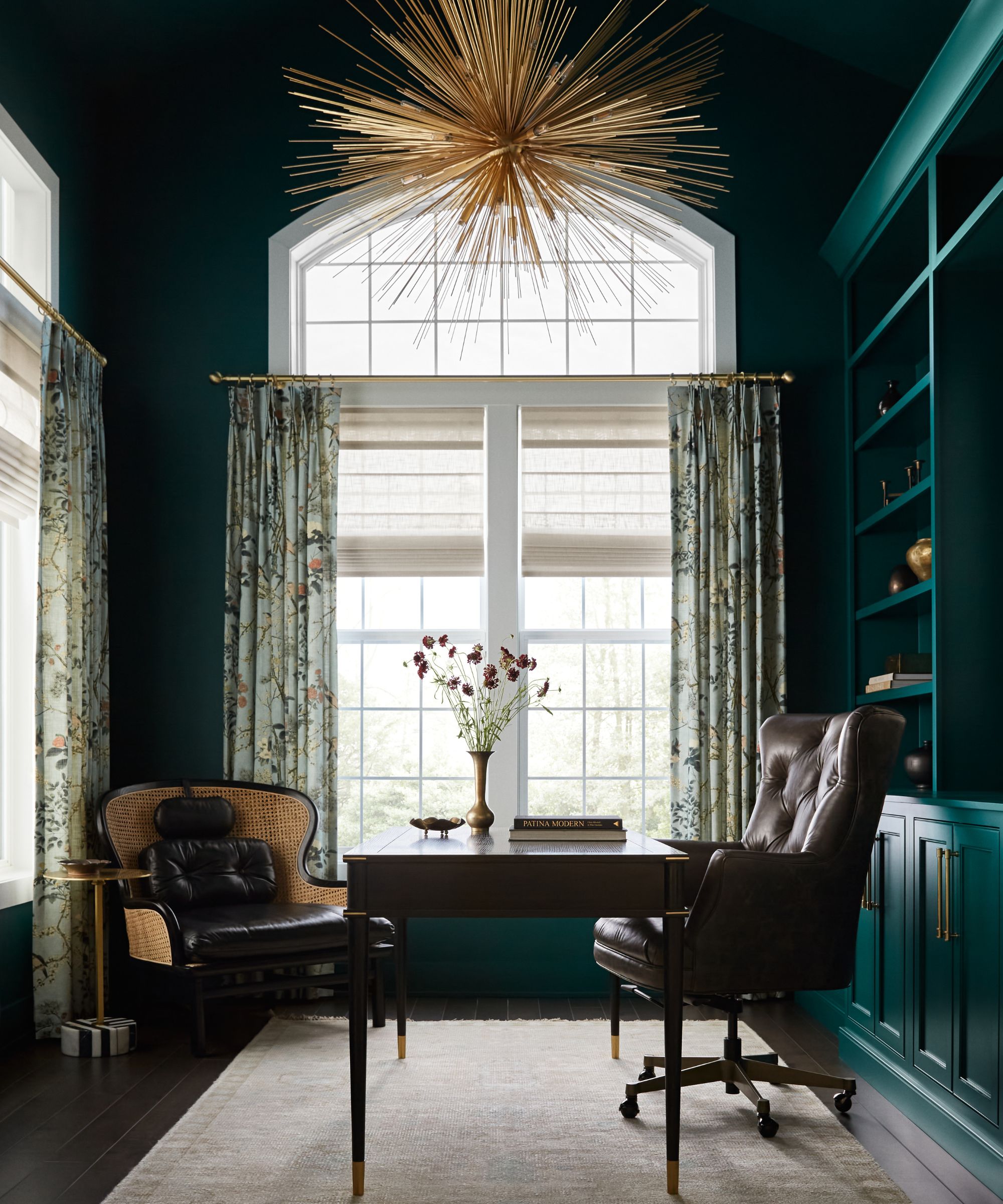 Home office with dark teal walls and bookshelf, large arched window with floral curtains and a gold star pendant light, a wooden desk, leather desk chair and rattan chair in the corner.