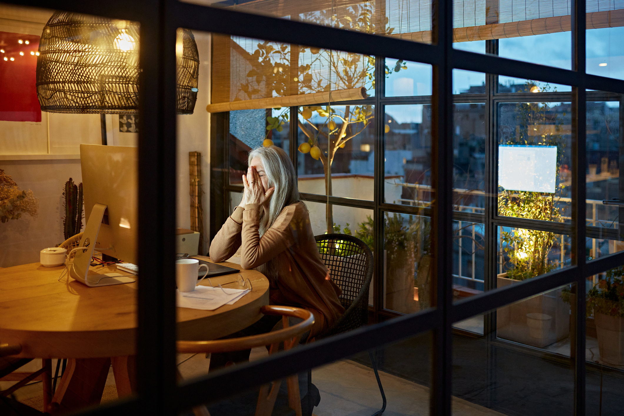 An older woman covers her face while looking at her computer. She is sitting in kitchen or home office in a glassed-in room.