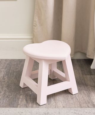 A light pink wooden heart stool, with a beige wall and curtain behind it on dark wooden flooring