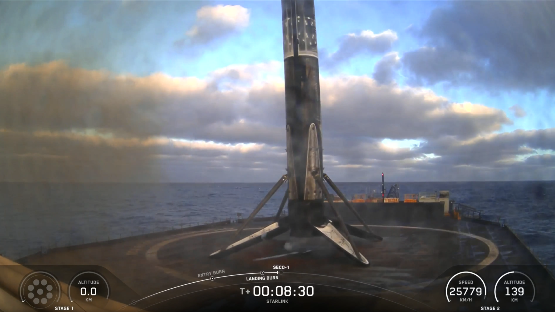 A charred rocket booster with four thin triangular lets stands on the flat black surface of a barge at sea.