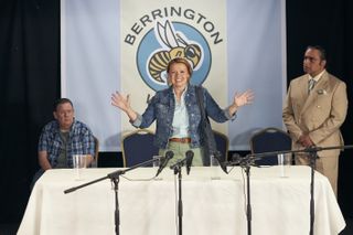 Gemma (Sian Gibson) stands in front of a Berrington Wasps logo at a table set up for a press conference. She has her arms outstretched and is smiling. Terry (Johnny Vegas) is sitting in one of the chairs to her right, and Gwyn (Sanjeev Bhaskar) is standing to her left looking alarmed