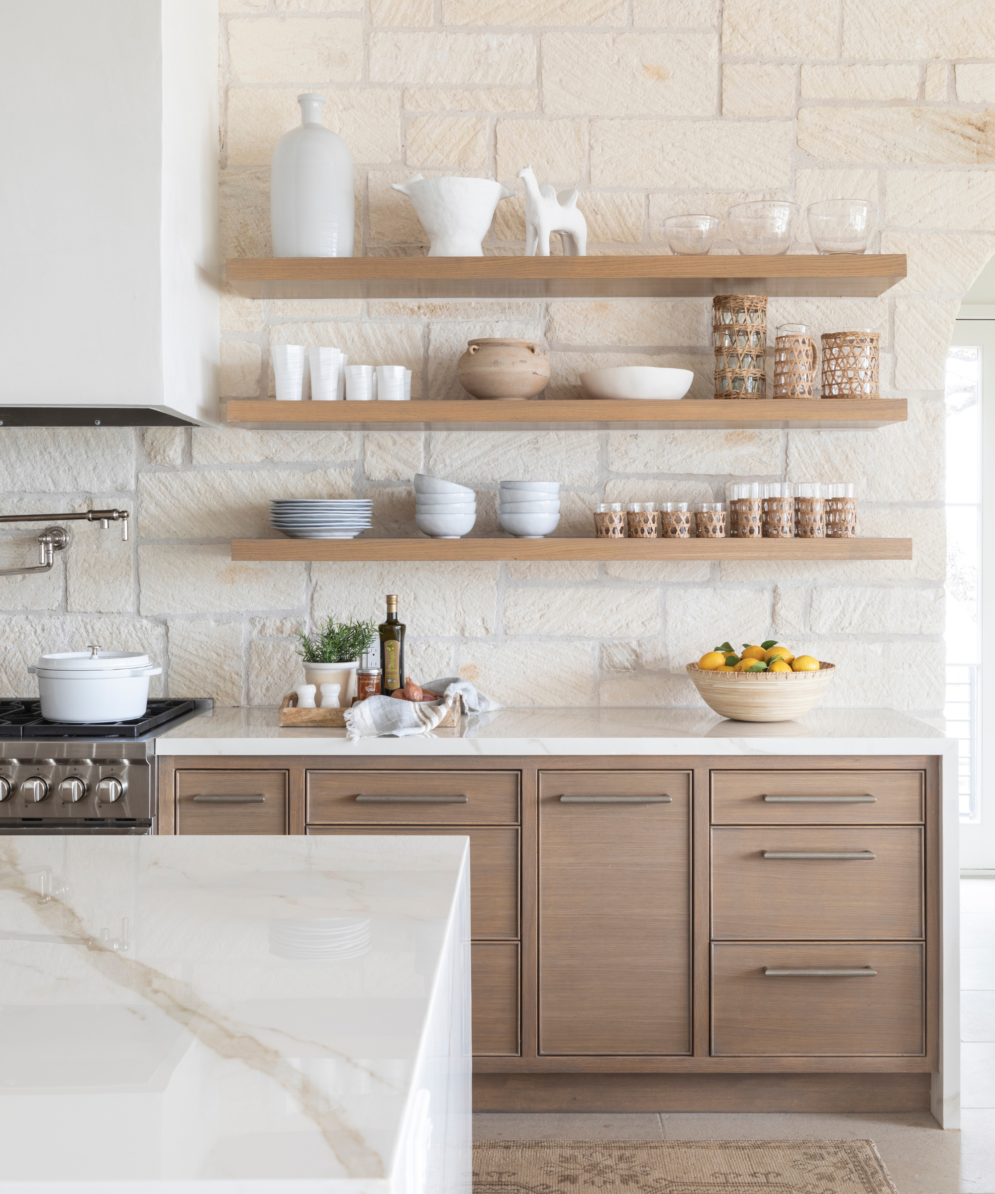 a kitchen with light wood cabinetry, white countertops, open shelves with cups and bowls, and an assortment of decor and kitchen necessities on the countertop