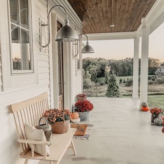 A white farmhouse porch with red and orange flowers in baskets, and two large wall mounted silver domed lamps