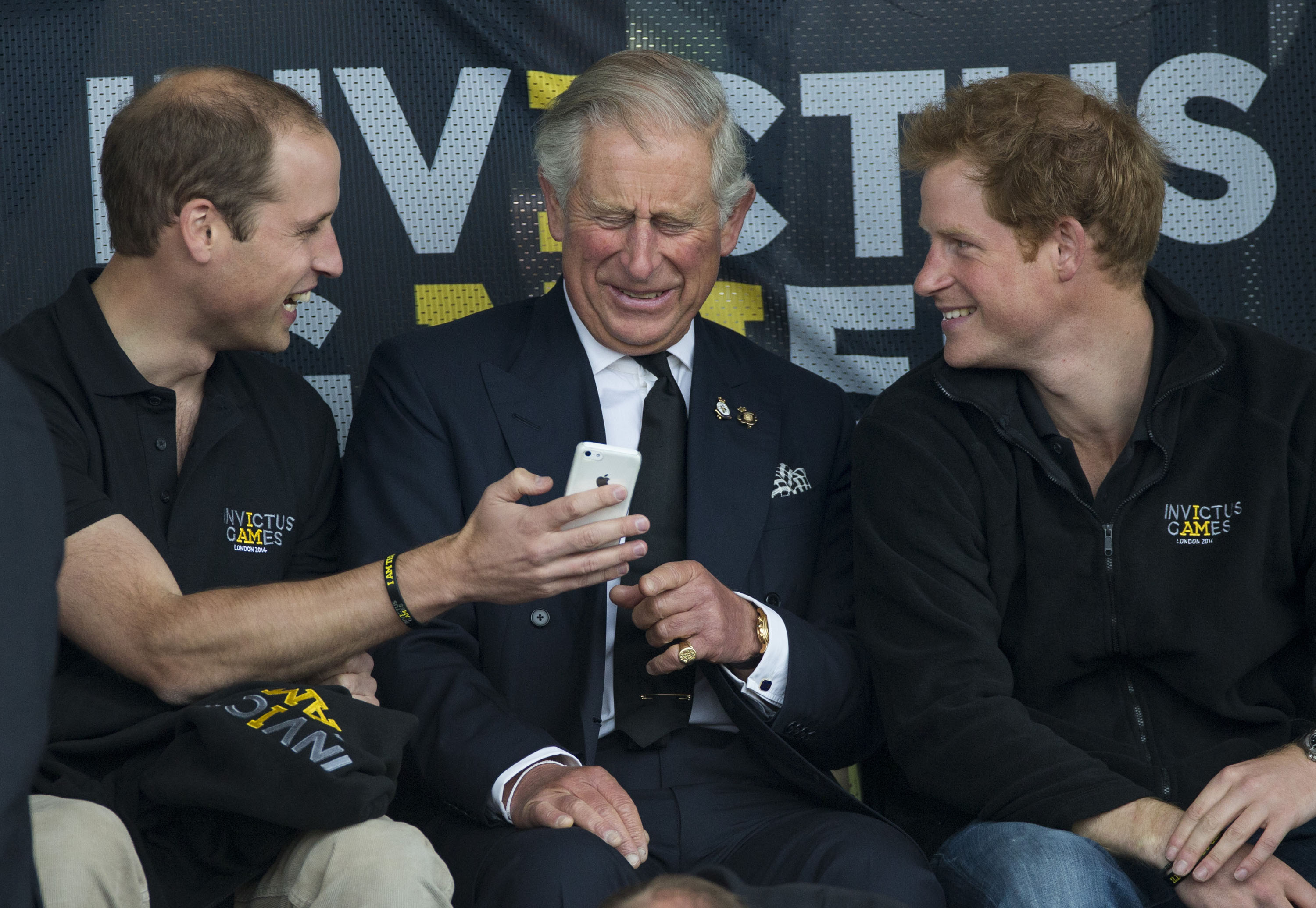 Prince William holding a phone up to King Charles and laughing next to Prince Harry