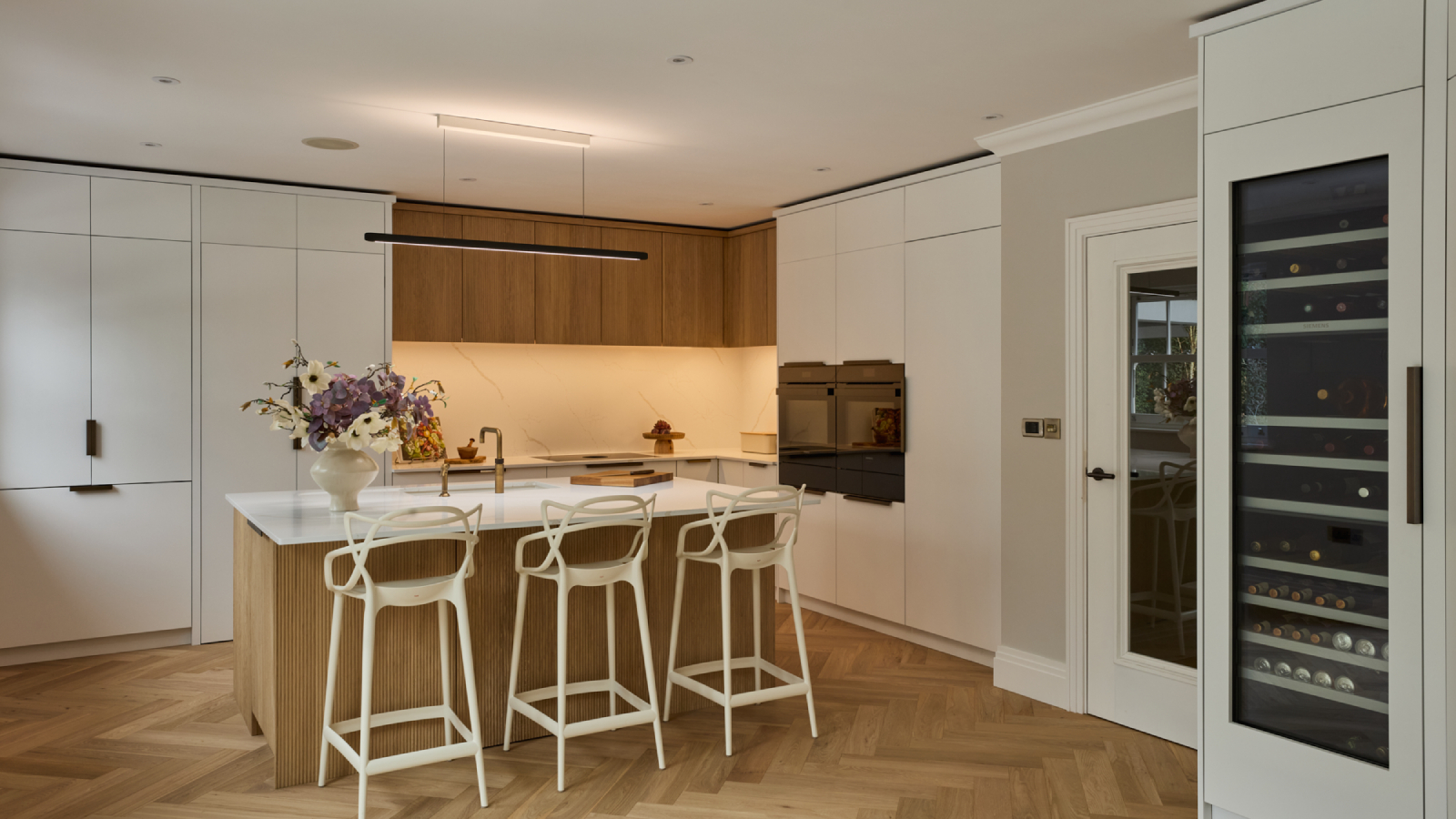 white kitchen with wooden detailing, kitchen island with bar stools, wine cabinet and herringbone wooden flooring