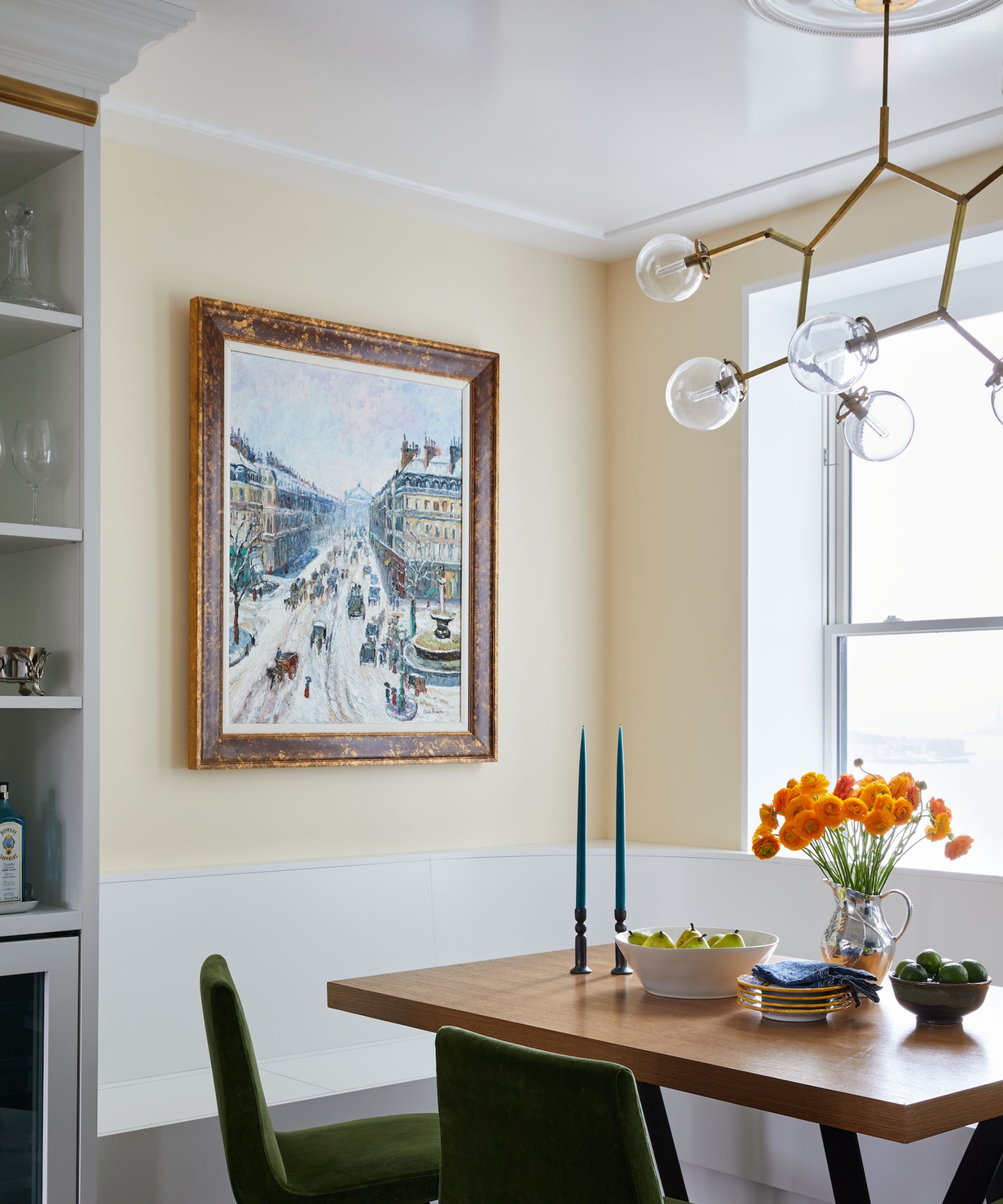 A breakfast nook with cream walls and a white lacquered ceiling. A large artwork on the wall, a wooden dining table, and a modern brass ceiling light fixture.