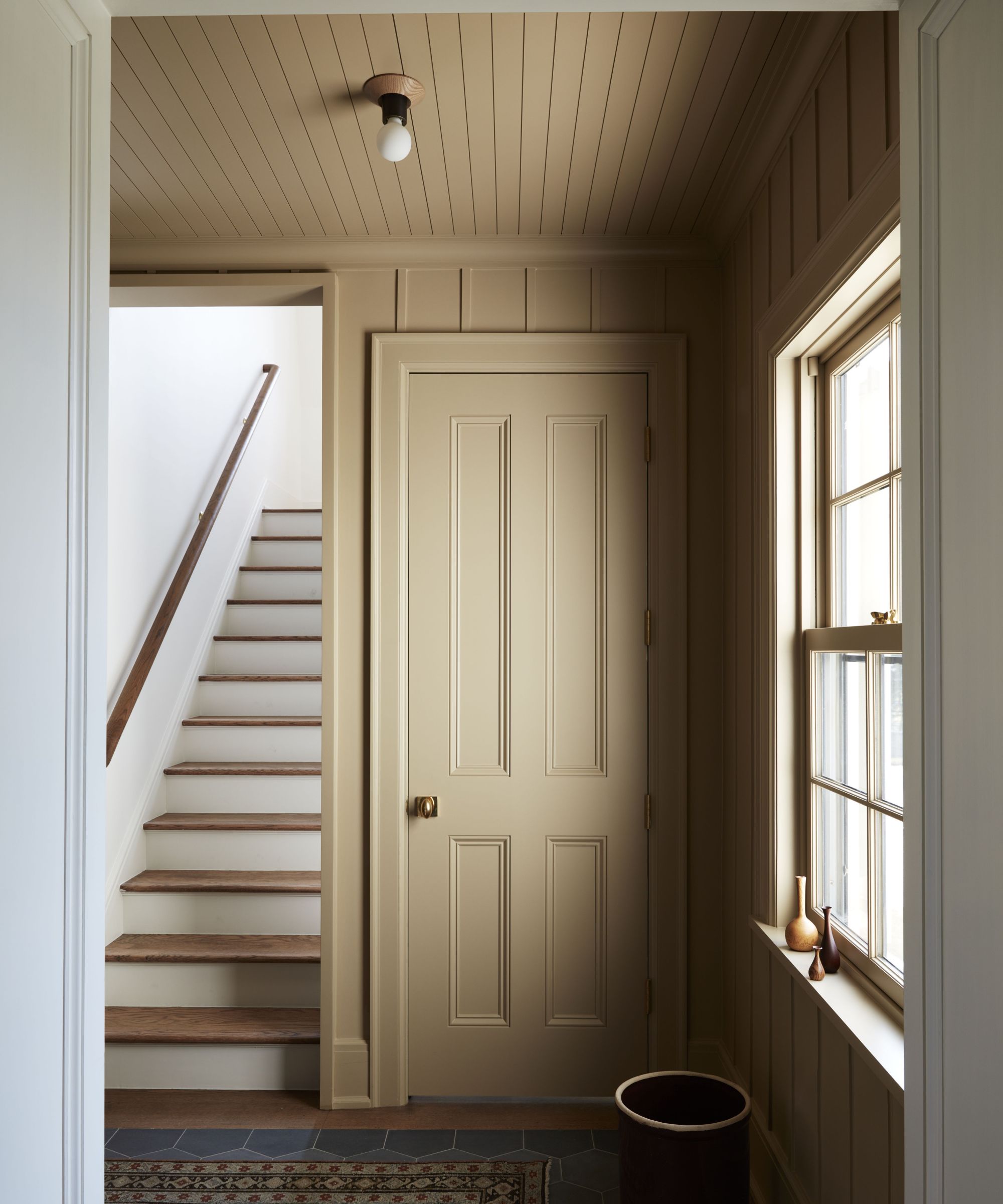 Entryway with a large window, shiplap ceiling and walls painted in caramel, and a door painted the same colour, with a wooden staircase to the left and white walls.