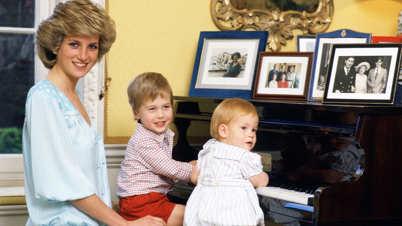 Princess Diana wearing a blue dress posing with baby Prince Harry and toddler Prince William sitting a piano covered with framed family photos