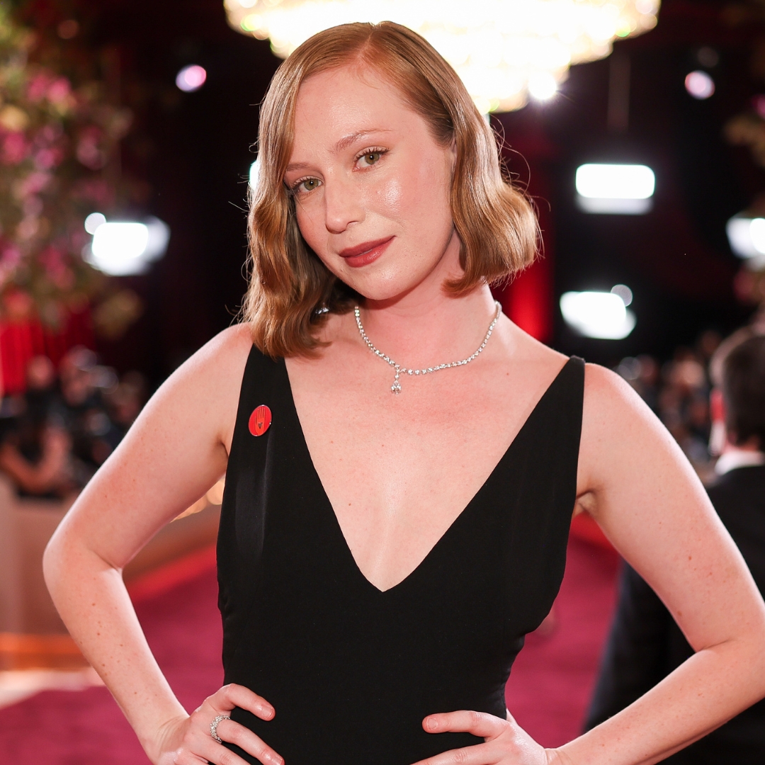 Hannah Einbinder pictured on the red carpet with wavy auburn bob tucked behind one ear and wearing a black V-neck dress