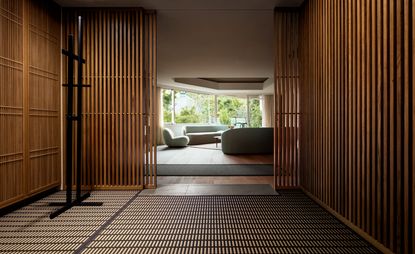 A wide hallway with floor to ceiling wood slats, leading to a step up into a lounge area with wide panoramic windows and large grey sofas and chair. 