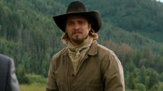 Luke Grimes as Kayce wearing a cowboy hat in Yellowstone. He&#039;s outside with mountains behind him. 