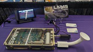An image of the LAMS system. It's a rectangular box with a clear sheath on top that has lots of technological instruments within. It's displayed on top of a table with a purple cloth.
