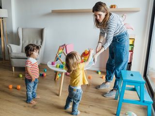 Babysitter in casual clothes playing with infant children in cozy playroom with colorful educational toys