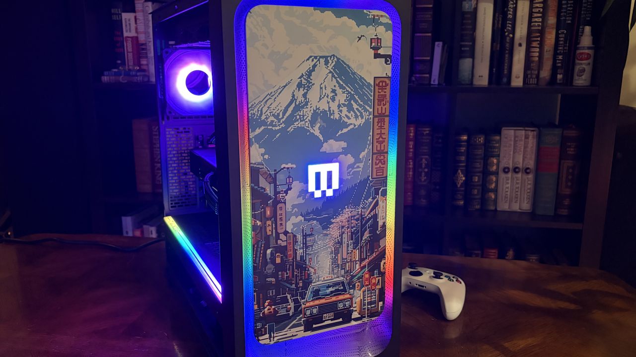 A Maingear MG-1 MK.II gaming PC with vibrant RGB lighting and a colorful case panel featuring pixel art of a cityscape with a mountain backdrop. A game controller rests nearby.