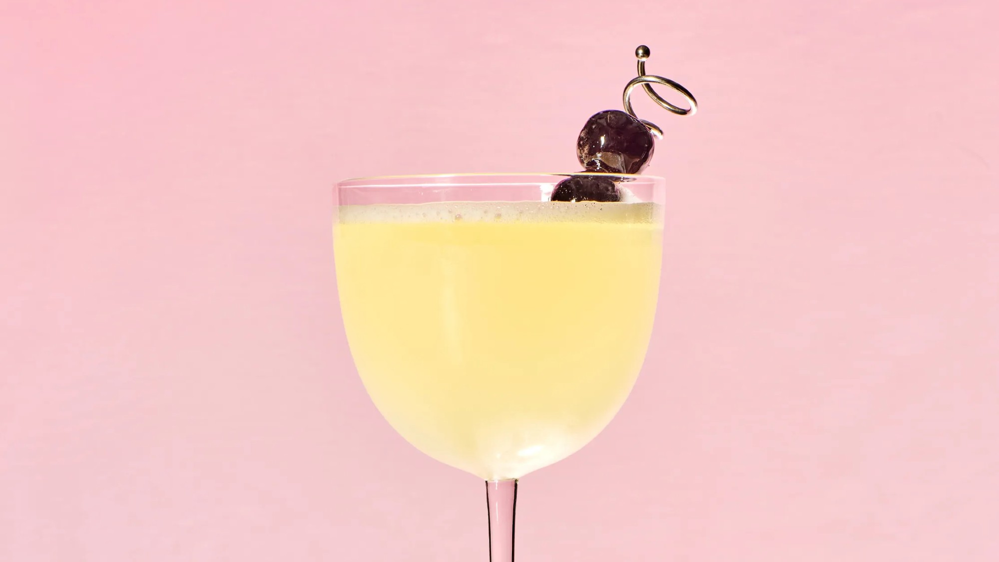 side shot of a yellow drink in a nick and nora glass with a cherry garnish on a skewer