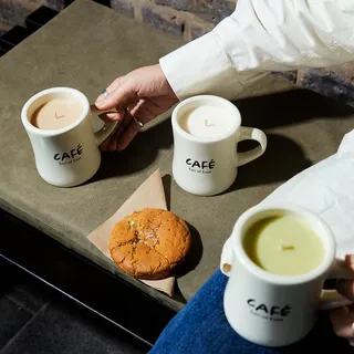 Three Earl of East-branded "mug"-shaped candles sit on a counter as a hand reaches for one of them as it stands next to an orange and sugar-coated cookie.