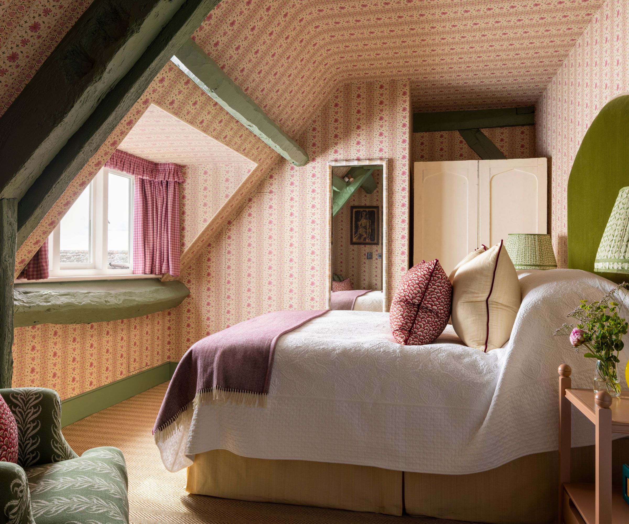 A traditional attic bedroom with pink wallpapered walls and ceiling, beams and woodwork painted green, a moss green headboard with white bedding and dark pink textiles.