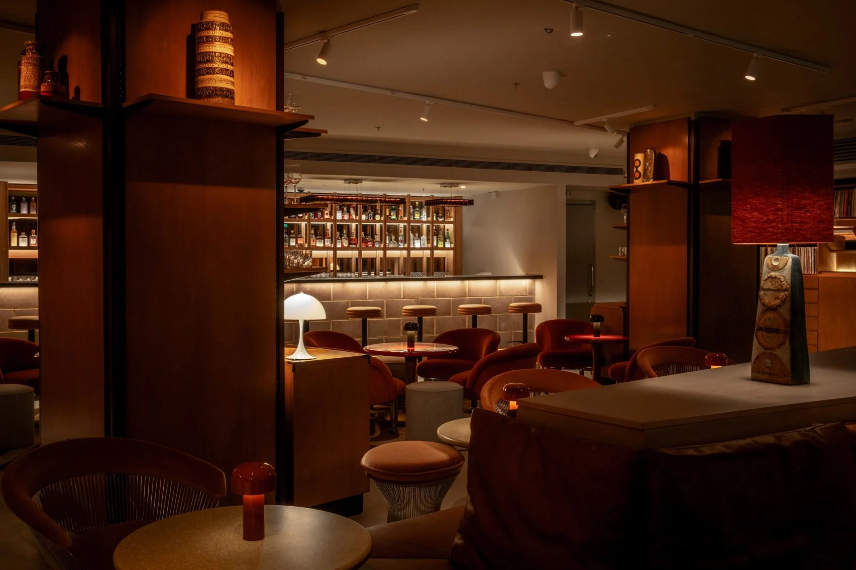 A suffused atmosphere absorbs a cocktail bar filled with 1970s sculptural furniture like mushroom shaped glass table lamps, leather mid-century stools, and a back-lit bar counter.